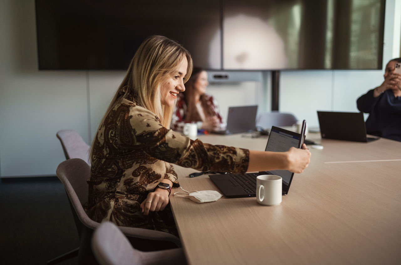 Een vrouw in een vergader ruimte achter een laptop met een kop thee of koffie
